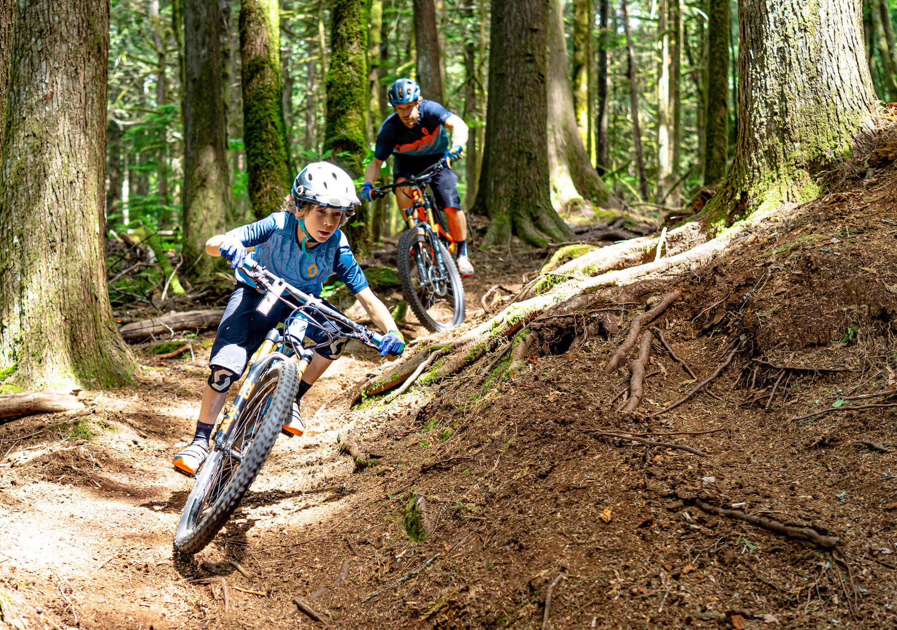 Two riders cornering through a root-covered forest trail